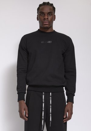 Man met gevlochten haar, gekleed in een zwarte trui en zwarte broek met witte tekst "LES HOMMES" op het shirt en trekkoorden, staand tegen een witte muur.