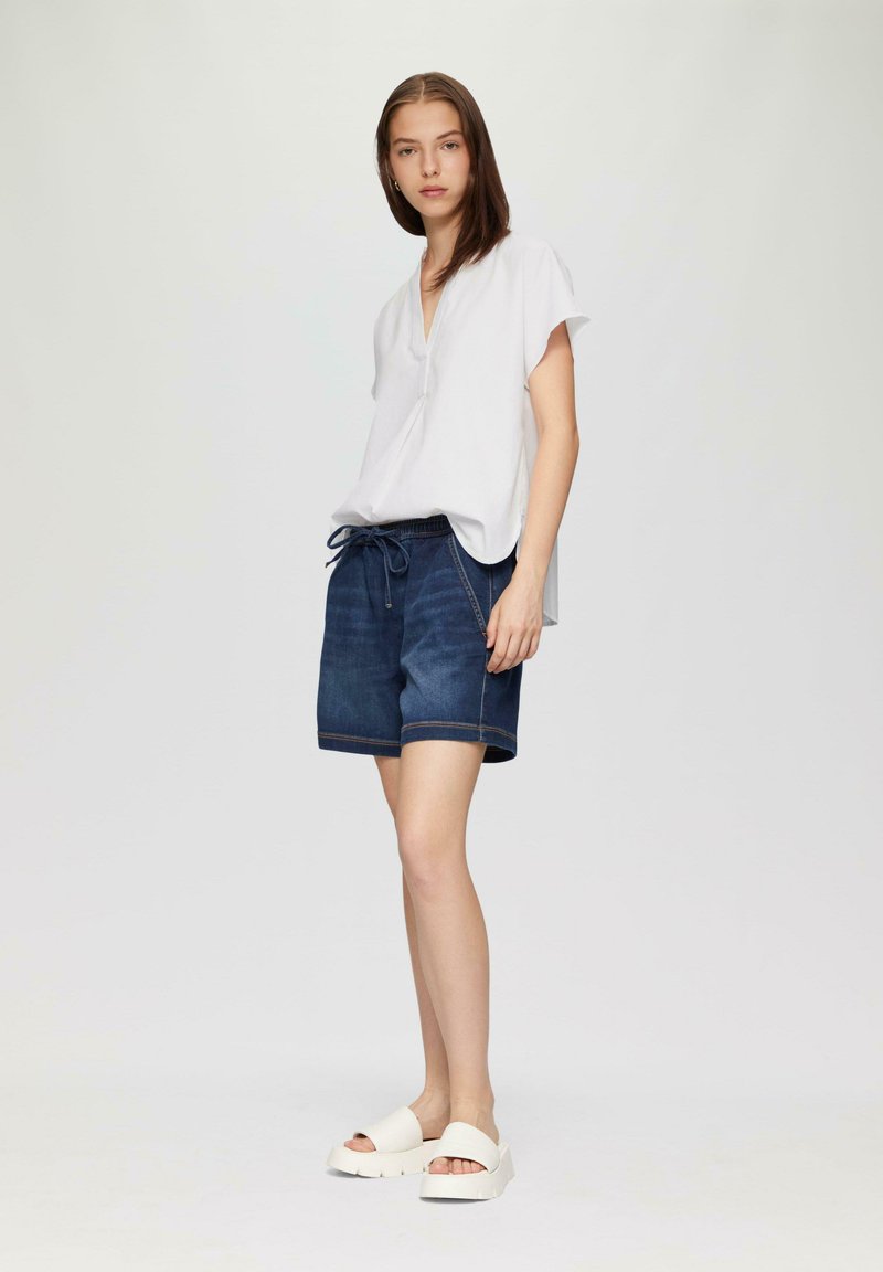 Witte top met korte mouwen en een V-hals, gecombineerd met donkere spijkerbroek shorts. De shorts hebben een elastische tailleband en een trekkoord, met zichtbare stiksels.