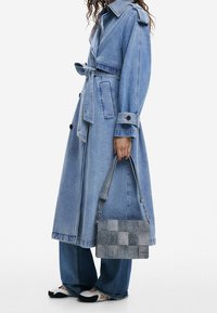 Denimowy płaszcz trench z paskiem w talii, zestawiony z szerokimi spodniami dżinsowymi. Niesie szaro-skalowy torbę. Czarno-białe buty uzupełniają stylizację.