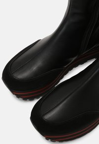 Stivaletti in pelle nera e camoscio con punta arrotondata, strisce rosse in contrasto sulla suola e suola in gomma testurizzata. Chiusura con zip laterale.