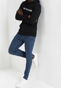 Sudadera negra con capucha y logo blanco de "Calvin Klein Jeans", combinada con jeans ajustados azules y zapatillas blancas, en una pose de pie.