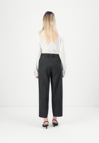 Personne avec de longs cheveux blonds portant une chemise blanche, un pantalon large sombre et des chaussures à talons noires, se tenant face à un fond blanc uni.