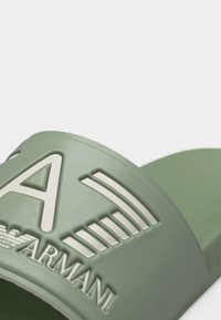 Σαγιονάρα slide σε απαλό πράσινο με ανάγλυφο λευκό λογότυπο "Armani" και στυλιζαρισμένο γράμμα "A" στο λουράκι, εμφανιζόμενη σε λευκό φόντο.