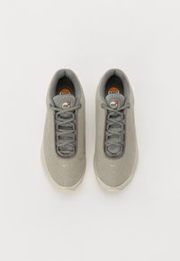 Szare siatkowe sneakersy o teksturowanej powierzchni, sznurowadła w ciemniejszym odcieniu szarości oraz okrągłe logo na języku. Podeszwa jest jasna i zaokrąglona.