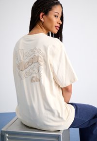 Beige oversized t-skjorte, korte ermer, har preget "ellesse"-logo i ulike størrelser på baksiden, laget av myk bomullsmateriale.