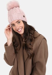 Pink knitted beanie with a large faux fur pompom, featuring a textured pattern and a small brown metal logo accent.
