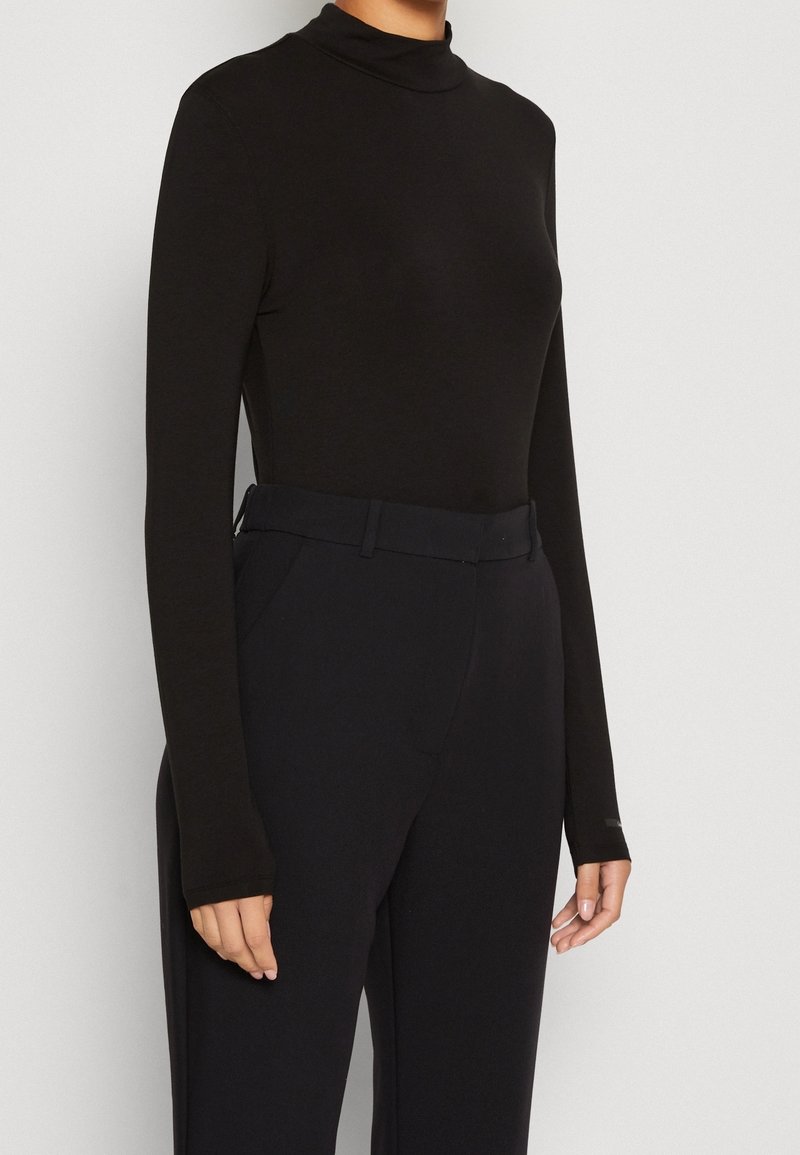 Schwarzes Turtleneck-Shirt mit langen Ärmeln aus glattem Stoff, taillierte Passform, kombiniert mit hochgeschnittenen schwarzen Hosen und einem passenden Gürtel.