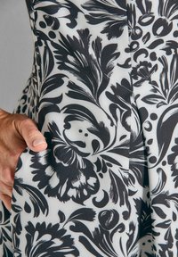 Hand rustend in de zak van een kledingstuk met zwart-wit bloemmotief tegen een neutrale achtergrond.