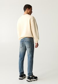 Lichtbeige sweater met ronde hals, blauwe vervaagde jeans met achterzakken, zwart-witte sneakers, effen achtergrond.