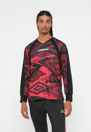 Ung mann med flettehår iført en rød og svart mønstrete Umbro langermet sportsgenser og svarte Umbro treningsbukser.