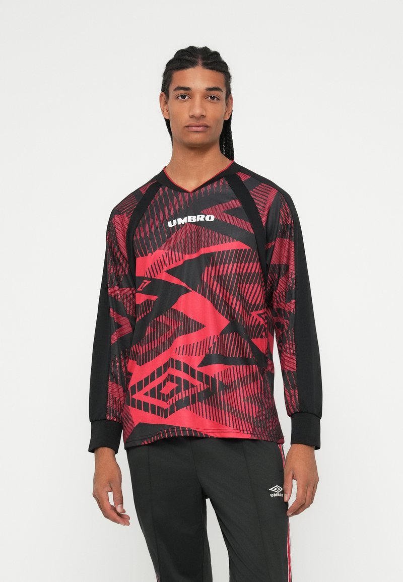 Fiatal férfi, aki fonott hajú, piros-fekete mintás Umbro hosszúujjú sportmezt és fekete Umbro melegítőt visel.