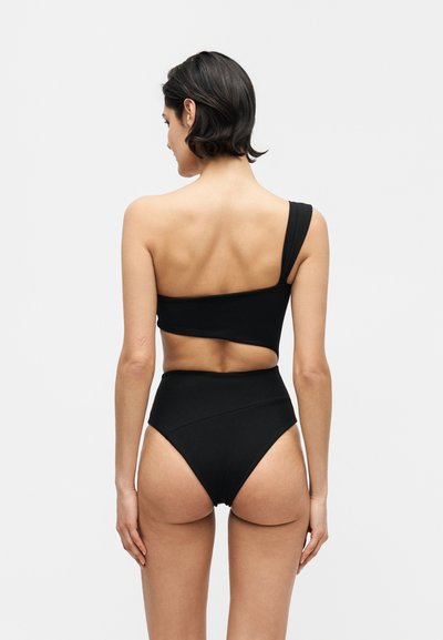 Γυναίκα με κοντά σκούρα μαλλιά που φοράει μαύρο ασύμμετρο τοπ μπικίνι με έναν ώμο και ψηλόμεσο κάτω μέρος μπικίνι, εμφανίζεται από πίσω.