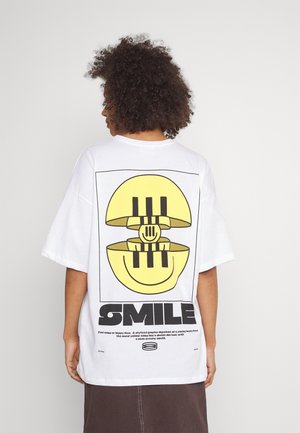 Personne aux cheveux bouclés de dos, portant un t-shirt blanc oversize avec un graphique jaune stylisé d'un visage souriant et un texte "SMILE" en gras dans le dos.