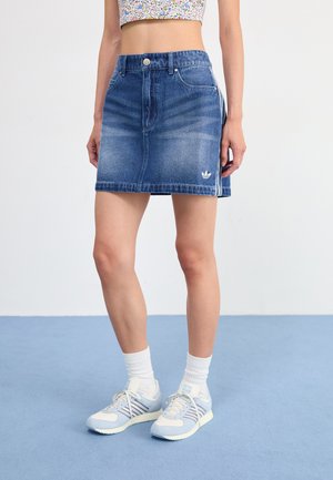 Denim skirt in medium blue with a fitted waist, front pockets, and a small white logo. Styled with light blue athletic shoes and white socks.