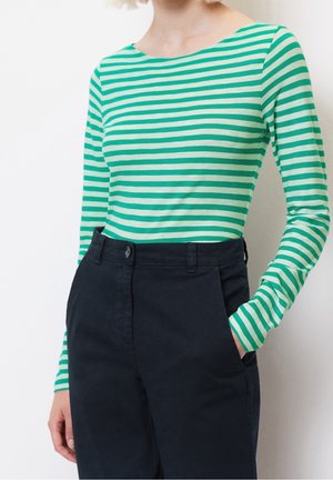 Vrouw met een groen-wit gestreept shirt met lange mouwen, ingestopt in een hooggetailleerde zwarte broek, met één hand in de zak.