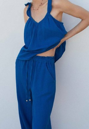 Personne portant un haut sans manches bleu lâche et un pantalon à cordon assorti, ajustant le haut pour montrer son abdomen contre un fond uni.