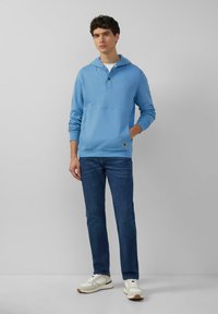 Lichtblauwe hoodie met een voorzak en knoopdetails, gecombineerd met donkerblauwe jeans en witte sneakers. Over het geheel een gladde textuur.
