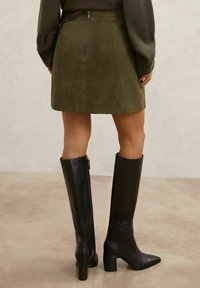 Fustă mini din piele întoarsă verde măsliniu, cu fermoar la spate, asortată cu cizme negre din piele până la genunchi, cu toc gros și vârf ascuțit.