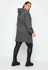 Oversized grå hoodieklänning med långa ärmar, framficka, ribbade muddar och dragsko i huvan, kombinerad med svarta ankelstövlar.