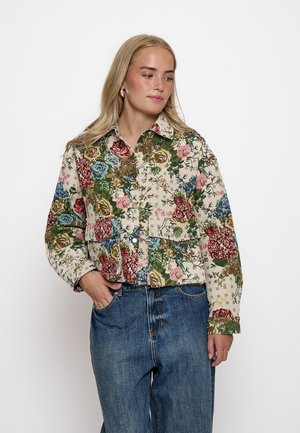 Mujer con cabello largo y rubio, que lleva grandes pendientes de aro, una chaqueta estampada con flores y vaqueros azules con una mano en el bolsillo.