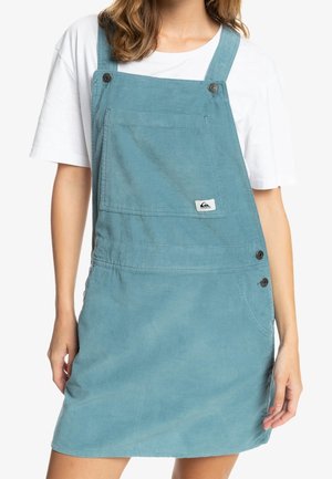 Blå kordflöjt overallklänning med en framficka, justerbara axelband och sidoknappar. Har en logotypdekal nära fickan.