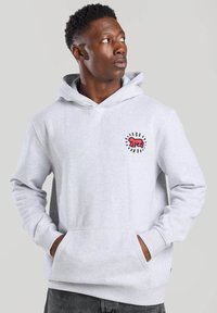 Gray hoodie with a kangaroo pocket, features a red embroidered logo surrounded by black lines on the left chest area, made of soft material.