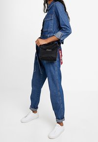 Czarna nylonowa torba na ramię z główną komorą na zamek błyskawiczny, uchwytem i ozdobnym paskiem. Noszona przez osobę w kombinezonie dżinsowym i białych sportowych butach.