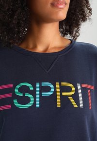 Primer plano de una mujer que lleva una sudadera azul marino con "ESPRIT" en letras de colores y a rayas en el pecho.