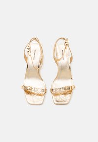 Sandales à talons hauts métalliques dorées avec un bout carré, des brides cheville et un accent boucle. Texture lisse et finition brillante.