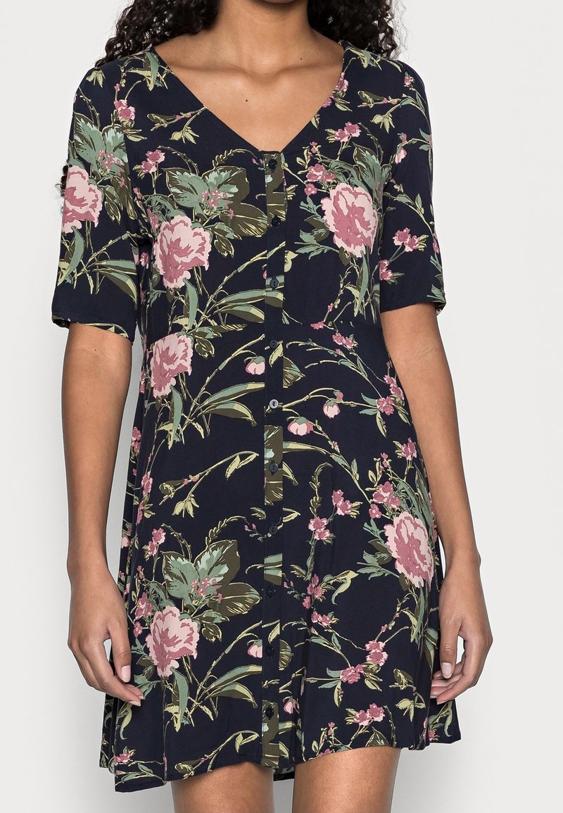 Abito floreale al ginocchio in tessuto blu navy, con maniche corte, scollatura a V e dettagli con bottoni lungo la parte anteriore. Accentuato da fiori rosa e verdi.