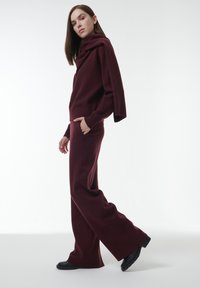 Bordeaux gebreide trui met een gedrapeerde kraag en bijpassende wijde broek, met textuurstof en zwarte platte schoenen. Minimalistisch ontwerp.