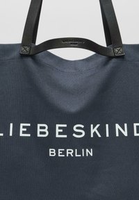 Granatowa torba na ramię z płótna z czarnymi skórzanymi paskami. Na przodzie umieszczono wyraźny biały napis „LIEBESKIND BERLIN”. Prosty, gładki design.