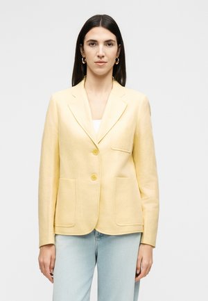 Vrouw met steil donker haar draagt een lichtgele blazer met twee knopen over een wit topje en lichtblauwe spijkerbroek, staand tegen een effen achtergrond.