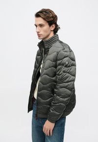 Επενδυμένο σκούρο πράσινο μπουφάν με ψηλό γιακά, υφή με ραφές που θυμίζουν κύματα, κλείσιμο με φερμουάρ και διακριτική σήμανση στο μανίκι.