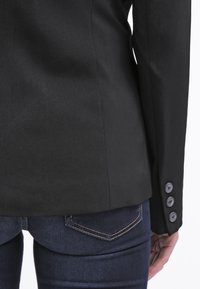 Veste noire sur mesure avec une texture lisse, dotée de cinq boutons sur la manche et d'une coupe structurée, associée à un jean en denim foncé.