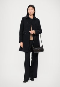 Palton dublu din lână închisă la culoare, cu guler, buzunare laterale și nasturi negri, asociat cu pantaloni negri cu picior larg și o mică geantă de umăr neagră.