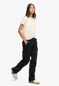 Zwarte cargobroek met zijzakken en een rode logo patch, gecombineerd met een plain wit t-shirt en witte schoenen met zwarte accenten.