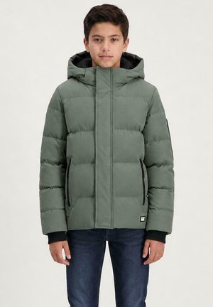 Jongen draagt een groene gewatteerde jas met capuchon en donkere spijkerbroek, staat voor een effen lichte achtergrond, kijkt naar voren.