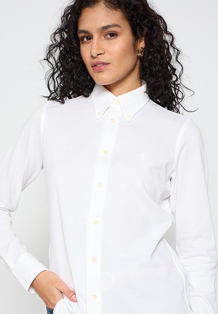 Camicetta bianca a maniche lunghe con colletto classico, dotata di bottoni in tonalità oro e un piccolo logo ricamato sul petto. Tessuto morbido.