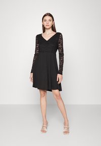 Vestido preto com corpete de rendas e mangas compridas, apresentando um decote em V e uma saia fluida que se alarga suavemente na barra.