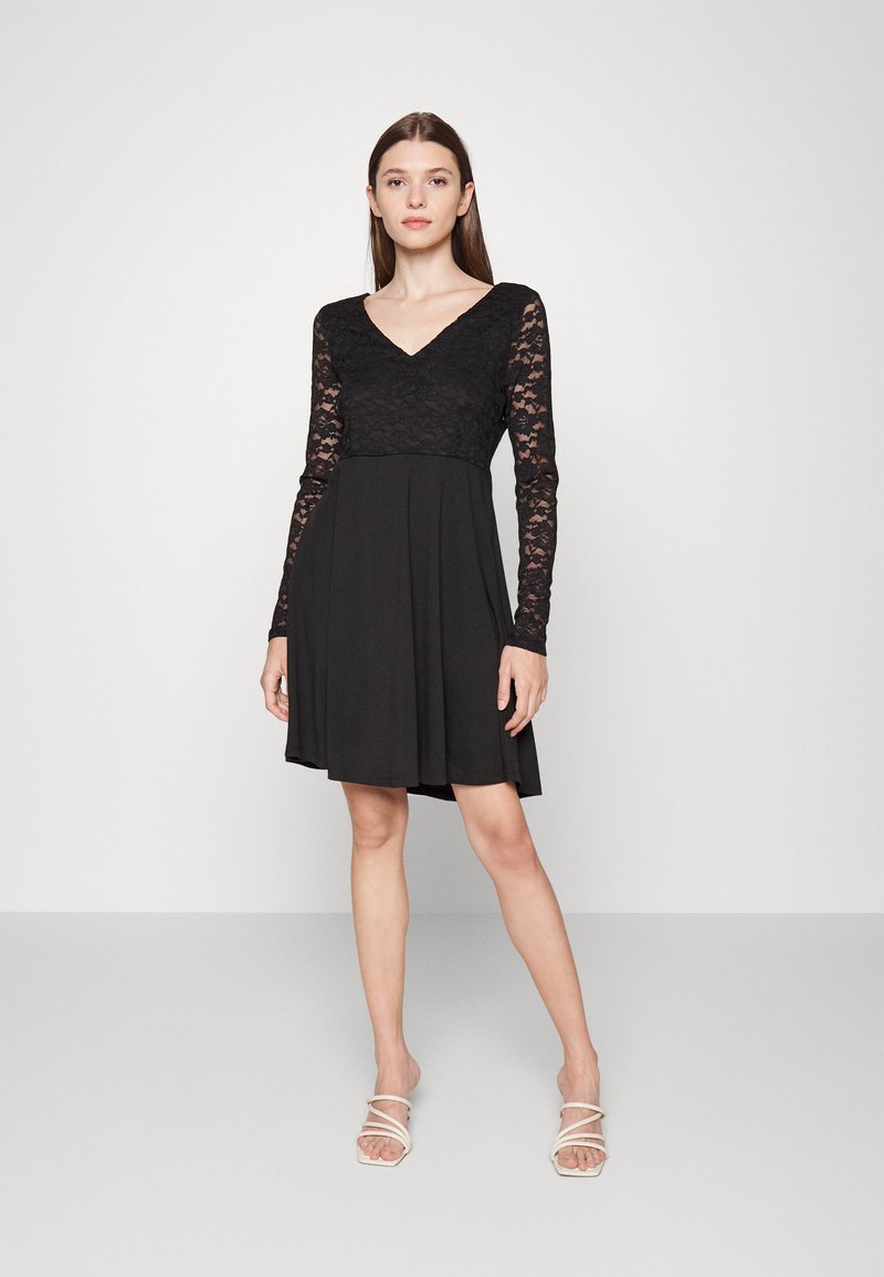 Vestido preto com corpete de rendas e mangas compridas, apresentando um decote em V e uma saia fluida que se alarga suavemente na barra.