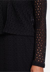 Schwarzes Spitzentop mit strukturiertem Muster aus offenen Webarten, langen Ärmeln und einem Lagenabschluss. Der Stoff ist leicht und atmungsaktiv.