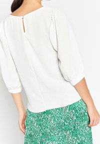 Vit blus med puffärmar, nyckelhålsdetalj på baksidan och mjuk textur, kombinerad med en grön mönstrad kjol med veck.