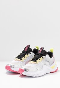 Baskets athlétiques blanches et grises avec des lacets noirs, des accents jaunes et des détails métalliques roses sur la semelle ; empeigne en mesh pour la respirabilité.