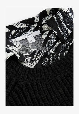 Sort strikket sweater med en mønstret krave i sort og hvid med blomsterdesigns. Inkluderer en dekorativ knap på kraven.