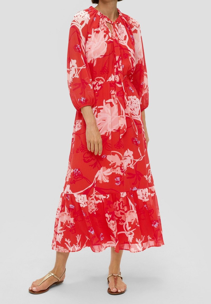 Femme portant une robe midi rouge à fleurs avec manches longues et ourlet à volants, associée à des sandales plates couleur camel, debout devant un fond uni.