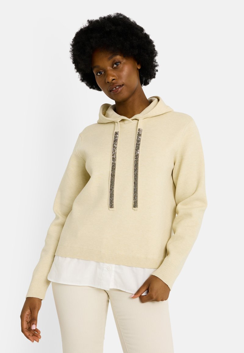 Sudadera beige con capucha y cordones plateados brillantes. Puños y dobladillo acanalados, llevada sobre una camisa blanca. Textura suave, corte holgado.