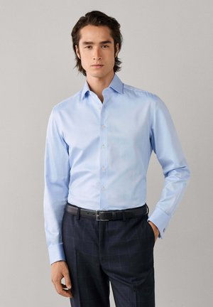 Man met halflang donker haar die een lichtblauw overhemd met knopen draagt, ingestopt in een donkere geruit broek met een zwarte riem.