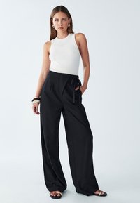 Zwarte wijde broek met hoge taille en een gestrikt ceintuur, gecombineerd met een aangesloten witte geribde tanktop en zwarte sandalen.