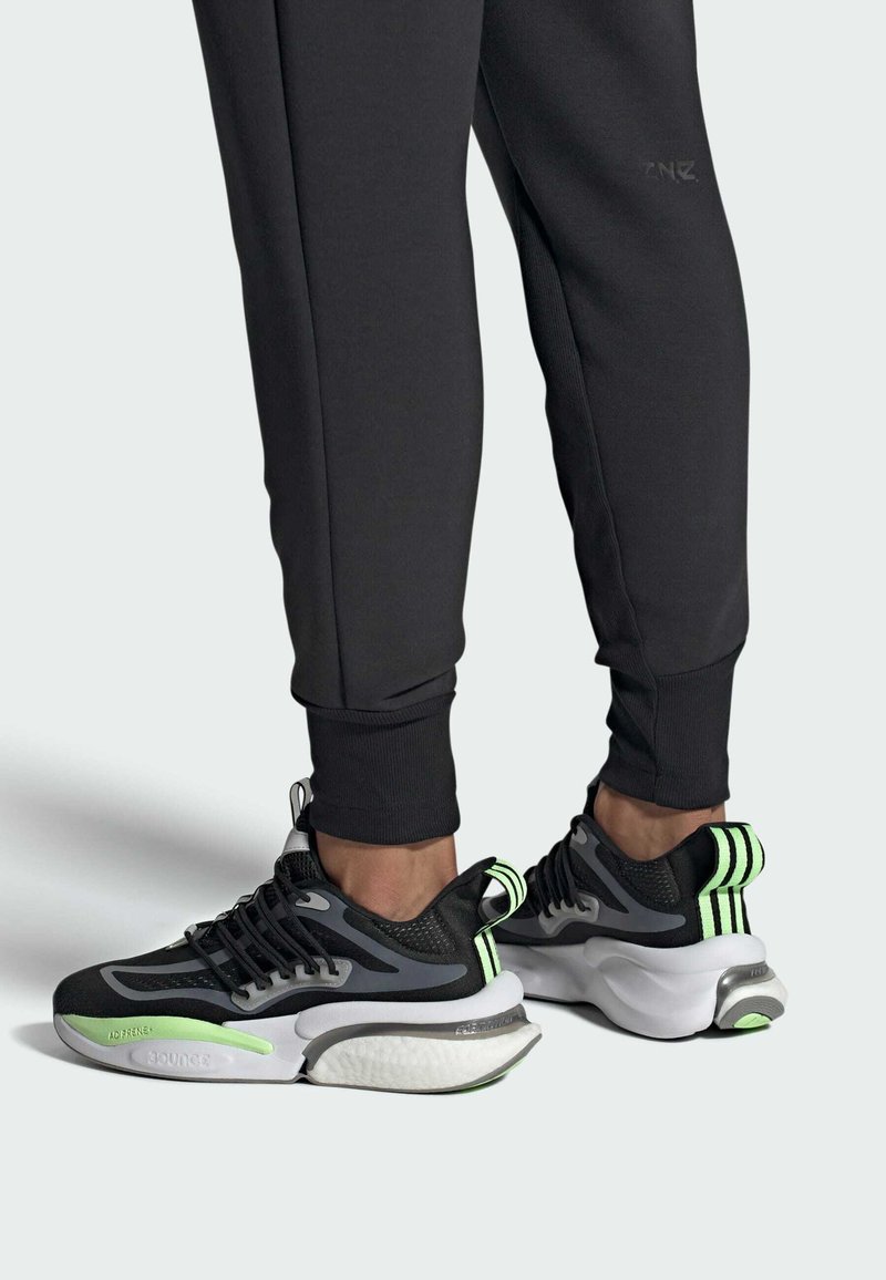 Chaussures de sport noires et grises avec des accents verts, tige en maille et synthétique, semelle épaisse avec amorti rebondissant et languette à l'arrière.